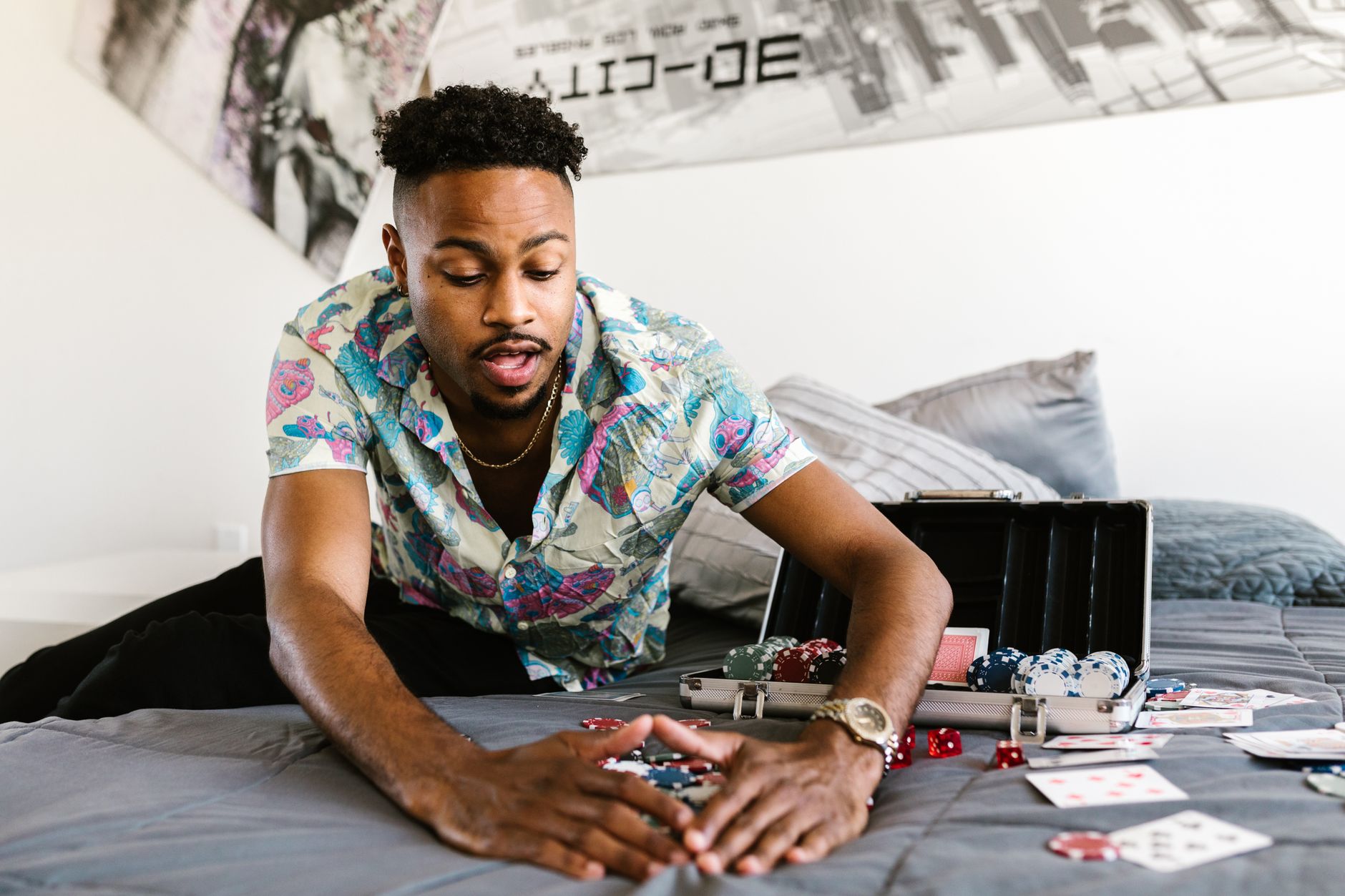 Homme en chemise colorée poussant des jetons de poker des deux mains sur un lit, près d’une mallette de jetons et de cartes éparpillées.