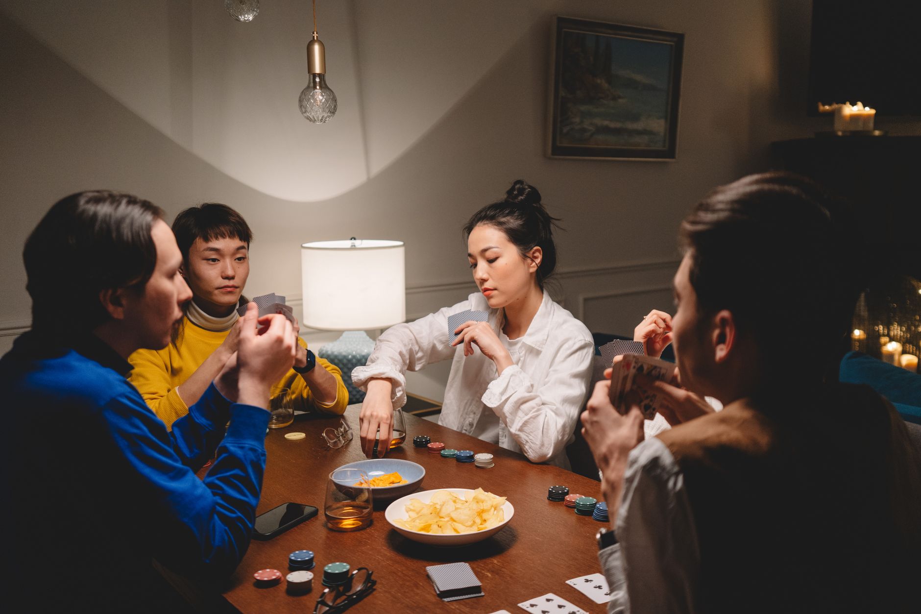 Quatre personnes jouent aux cartes autour d’une table en bois avec des jetons de poker, un bol de chips et un verre, dans une pièce à la lumière douce.